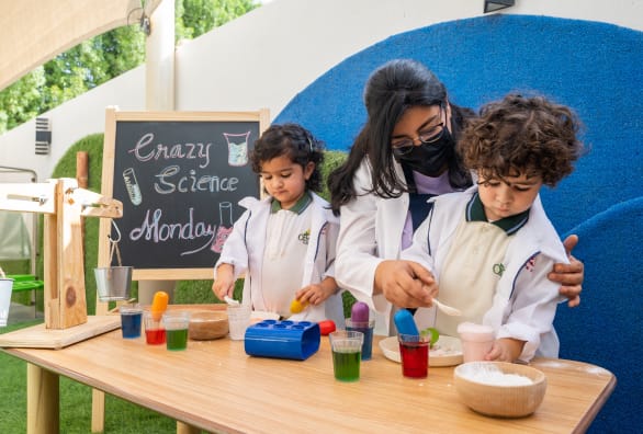 A woman in a lab coat with children in front of a chalkboard.